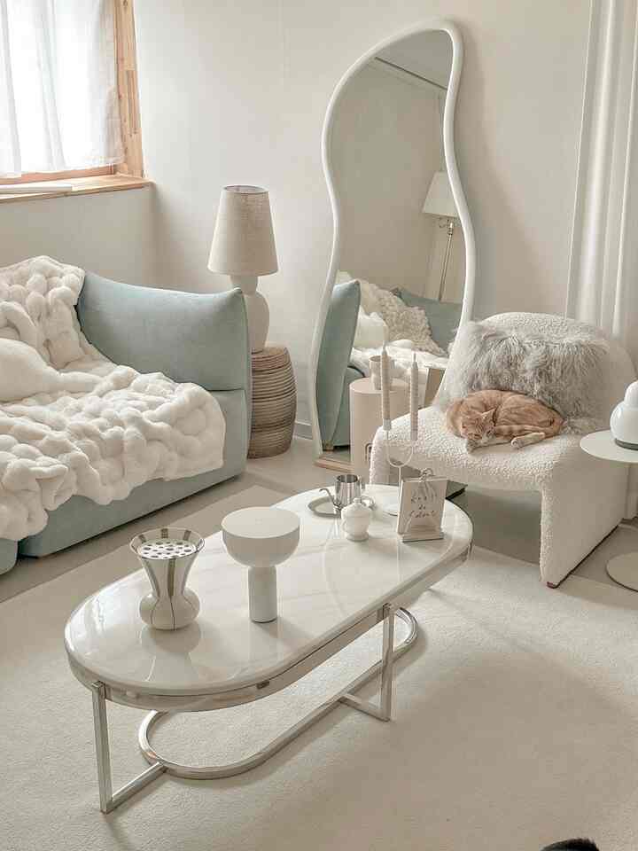 Beige and ivory toned living room featuring soft sofa, armchair with cat, wavy mirror, and warm lighting creating a cozy space