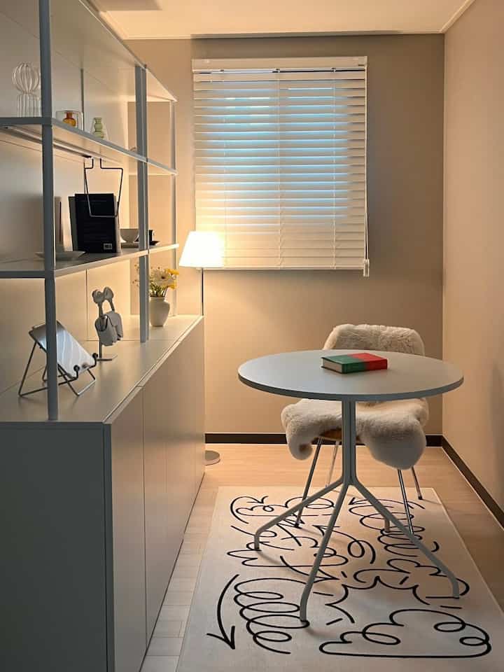 A white and beige toned walk-in closet featuring a round table, cozy chair with soft cover, lamp, and patterned rug creating a warm home cafe atmosphere