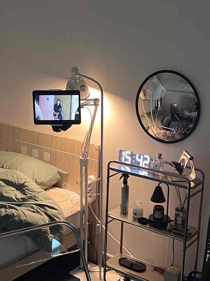 ナチュラルトーンとメタルラックが調和するワンルーム寝室、スマートデバイスホルダーとモダンな照明が配置された印象的な空間