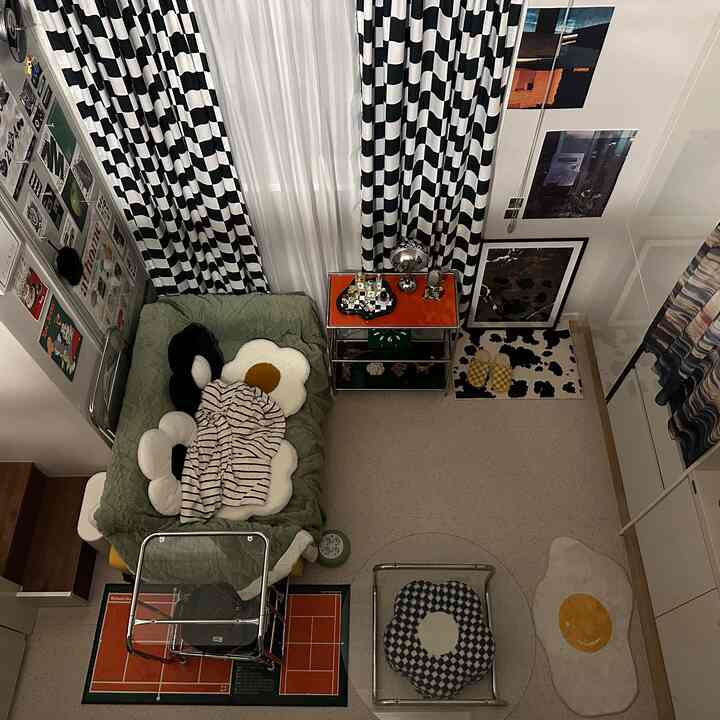 白黒のチェック柄カーテンと緑色の毛布が敷かれたワンルーム、バスケットトロリーと花の形のクッションが配置された印象的な空間