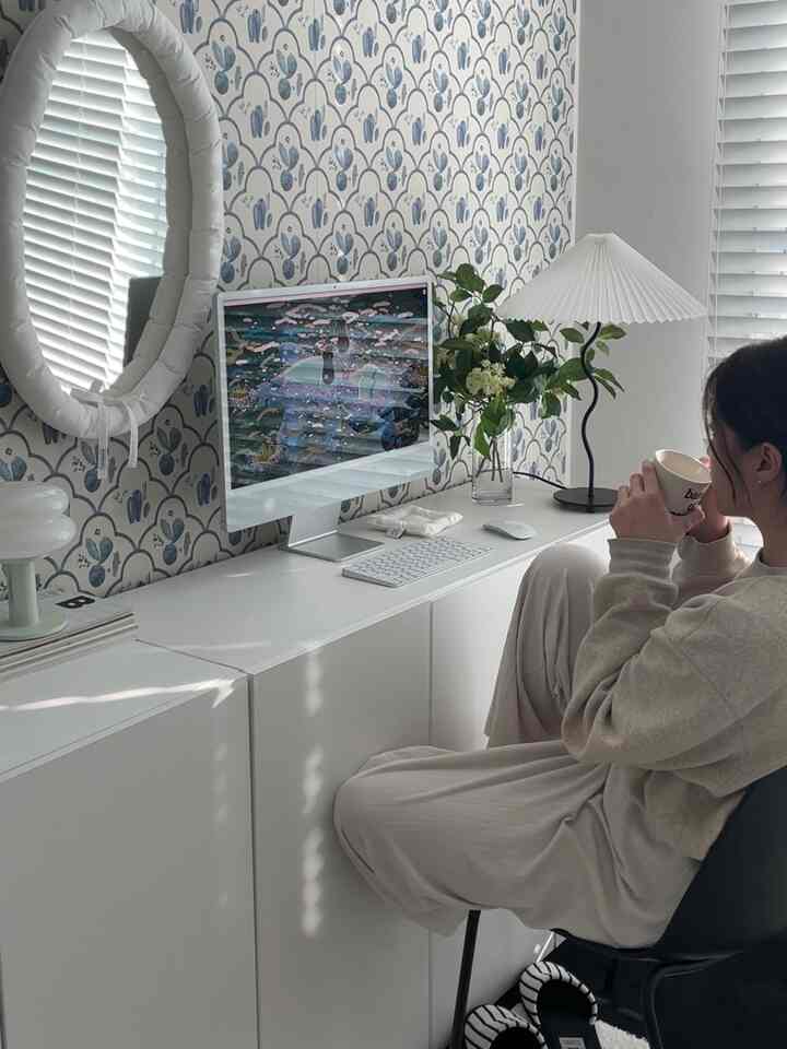 White and blue toned home office space featuring a desk with an iMac and table lamp, round mirror on patterned wall in a modern interior