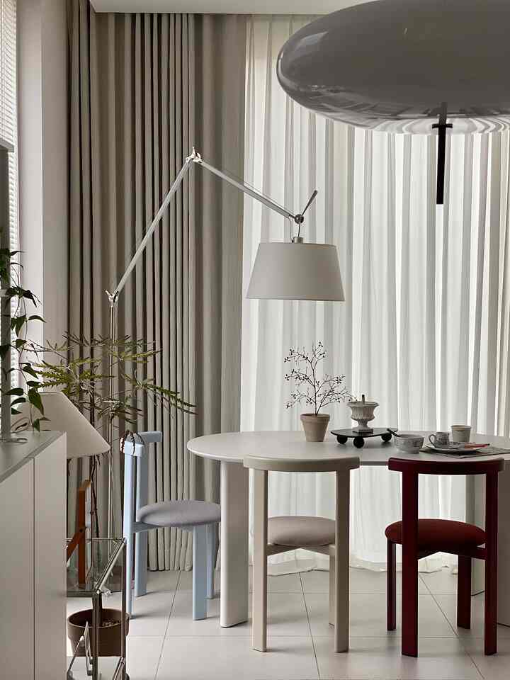 A living and dining room in white and beige tones featuring a round table and colorful chairs, with modern, soft curved lighting creating a cozy atmosphere