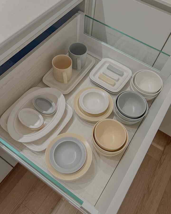 パステルカラー調のキッチンの引き出しの中に、多様なサイズのベージュとアイボリーの食器セットが整頓された印象的な空間