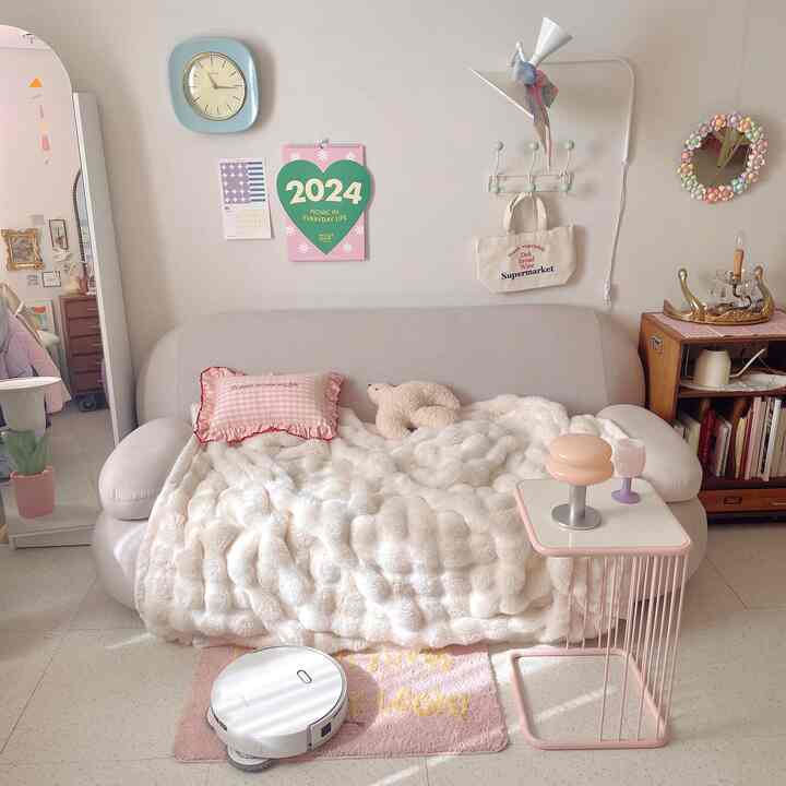 柔らかいホワイトとピンクの色調のリビング、グレーのソファとふわふわのブランケット、サイドテーブルと壁掛け時計が配置された雰囲気のあるインテリア