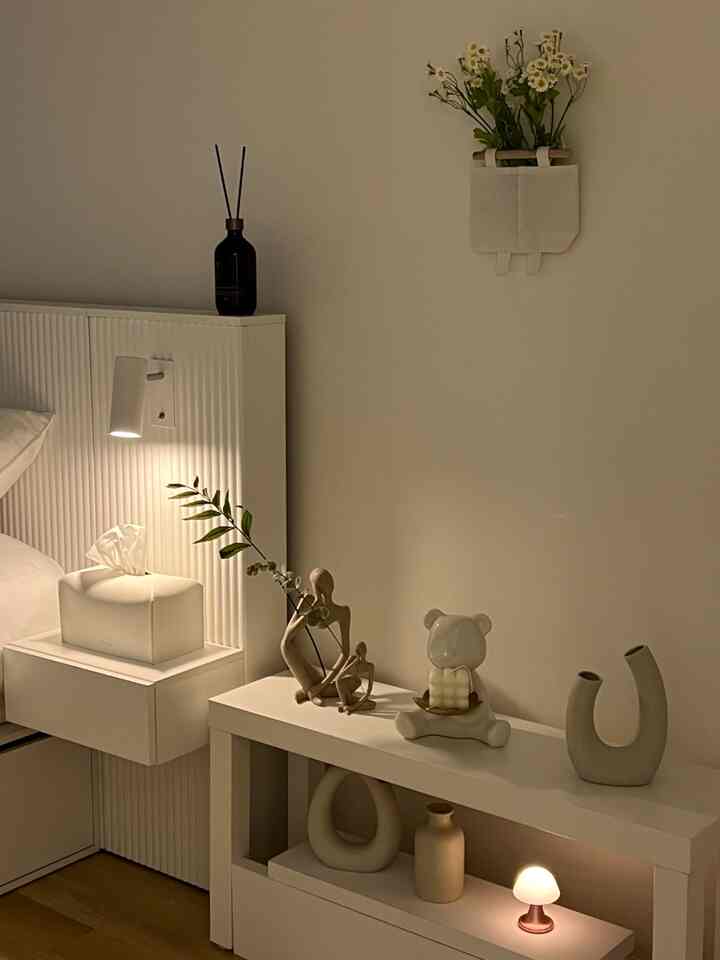 ホワイトトーンの寝室空間、中央左寄りにベッドとナイトテーブル、右側にモダンなオブジェと柔らかな照明が配置された雰囲気のあるインテリア