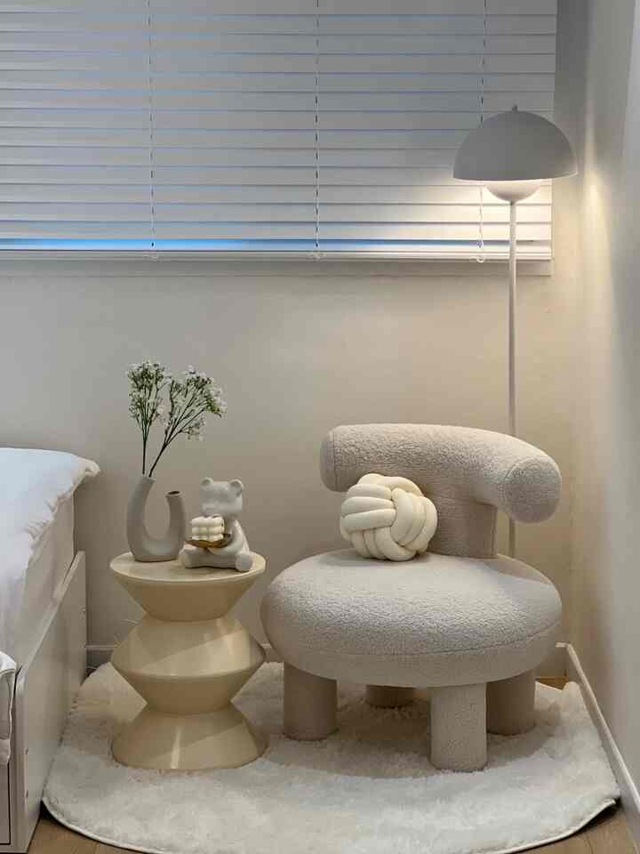 明るいホワイトトーンの寝室コーナー、丸みのあるベージュ色のスツールと柔らかな質感のソファ、フロアランプが配置された温かみのある空間