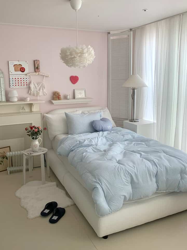 ピンクの壁とパステルブルーの寝具が配置された居心地の良い寝室、白いカーテンと柔らかな照明が雰囲気を演出している