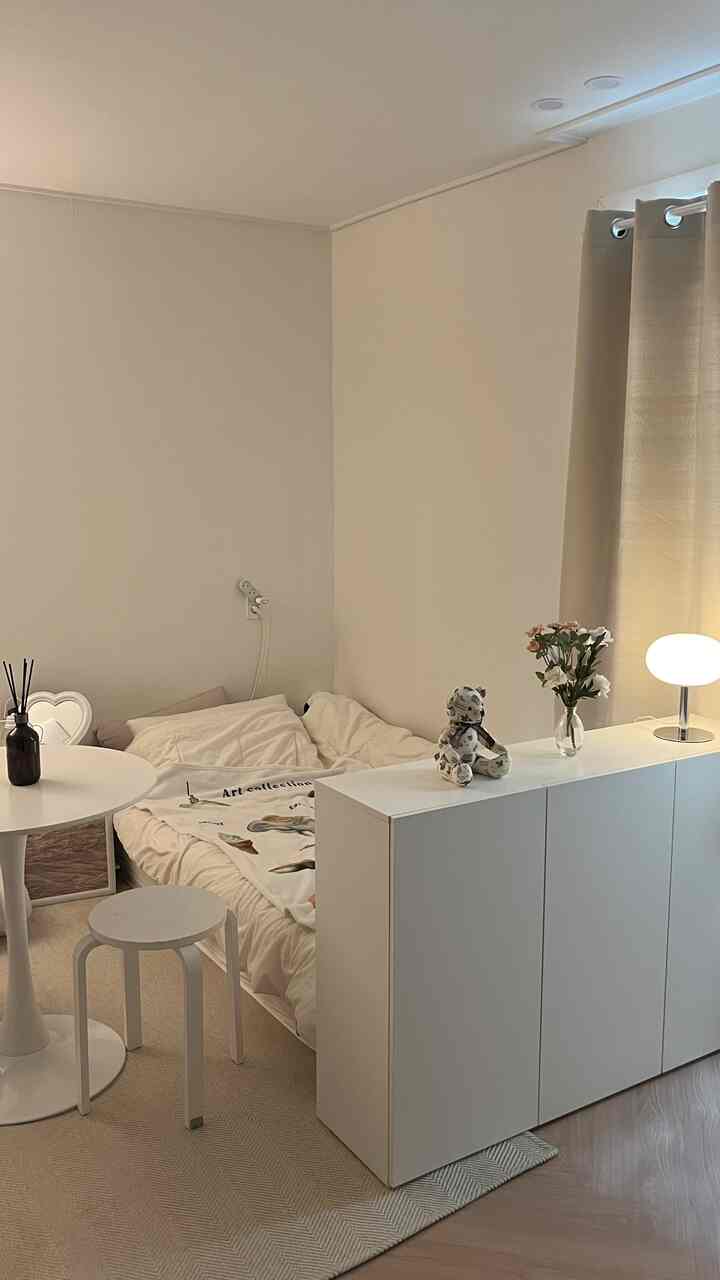 ホワイトとベージュのトーンが調和した小さなワンルーム寝室、円形ダイニングテーブルとスツール、シンプルで雰囲気のある照明のあるインテリア