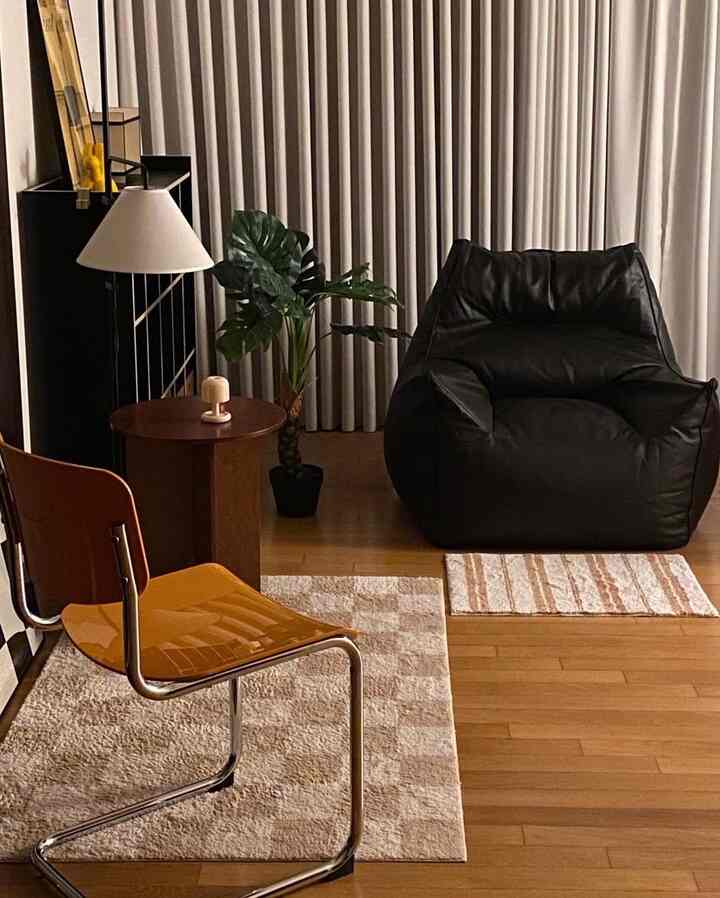 Natural and modern living room with warm wood-tone flooring, beige curtains, black bean bag chair, and orange stool