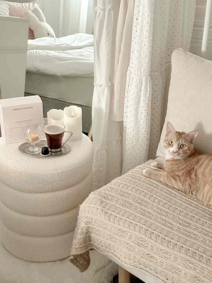 Beige and white toned bedroom featuring an armchair with a cat, round ottoman with LED candles creating a cozy atmosphere