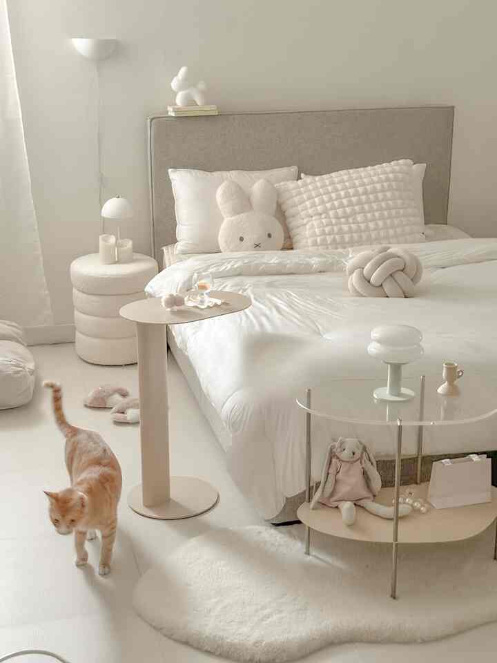 White and beige toned bedroom featuring soft bed, pet cat, baby stuffed toys, and storage tables in a natural modern style