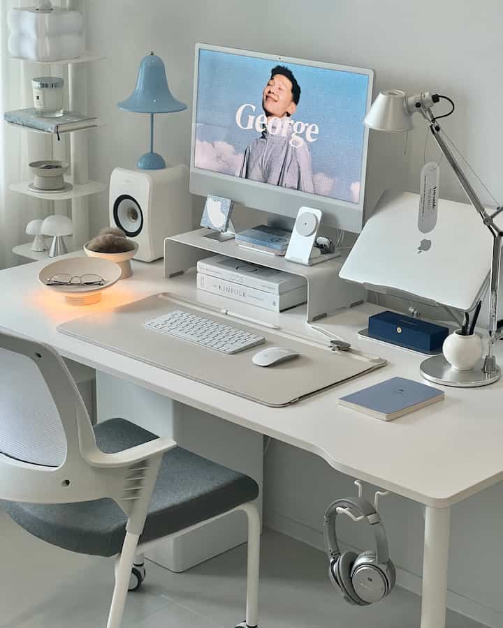 White-toned home office desk space featuring an iMac monitor on a monitor stand, table lamps, and an office chair in a clean workspace