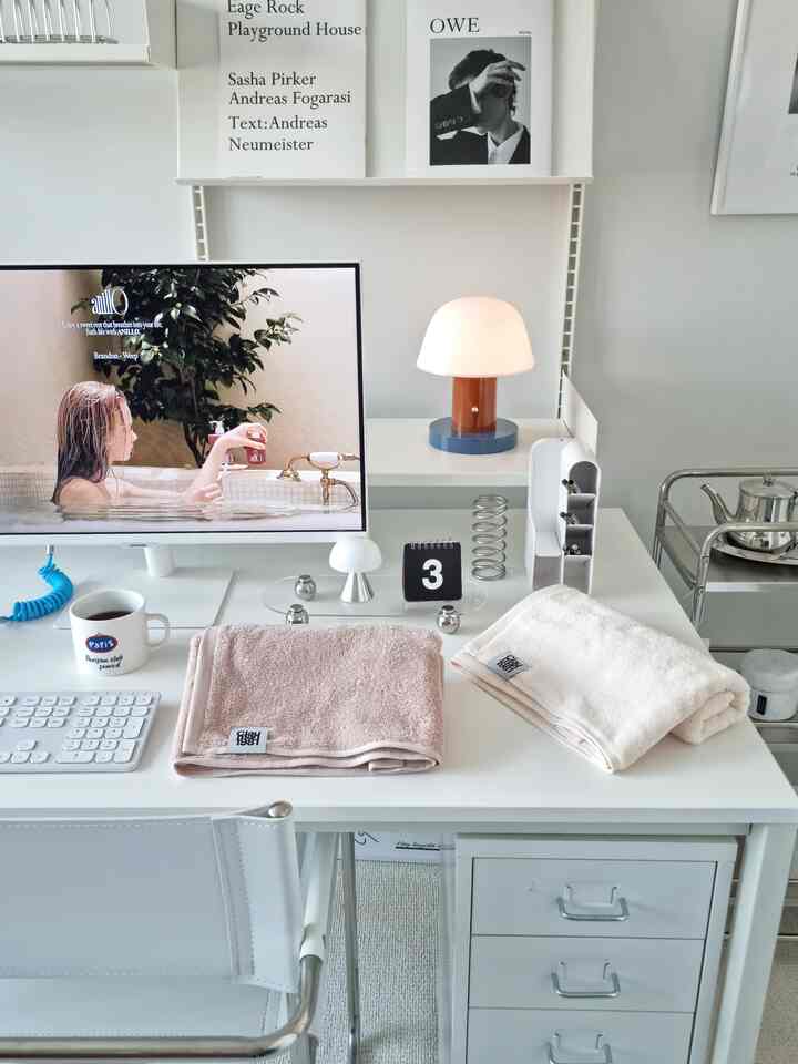 白とベージュのトーンのホームオフィスデスク、タオルとモニター、照明が配置された清潔でモダンな空間