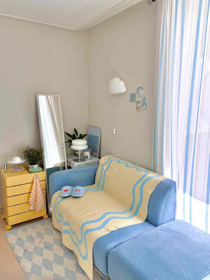 淡いナチュラルトーンの壁と床、青いソファと黄色のキャビネット、パターンカーテンがある居心地の良いリビング