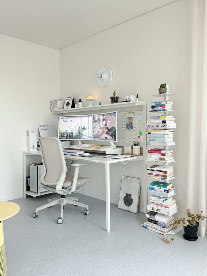Bright and airy white-toned study featuring a white desk and chair, dual monitors, bookshelf, and plants in a neat home office setting