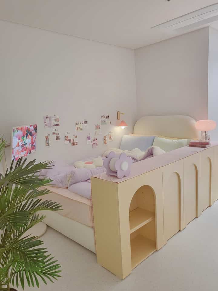 ベージュとアイボリーの色調の寝室、中央にベッドと収納キャビネットが配置され、壁には小さな写真カードが貼られ、左下に緑の植物が置かれた雰囲気のあるインテリア