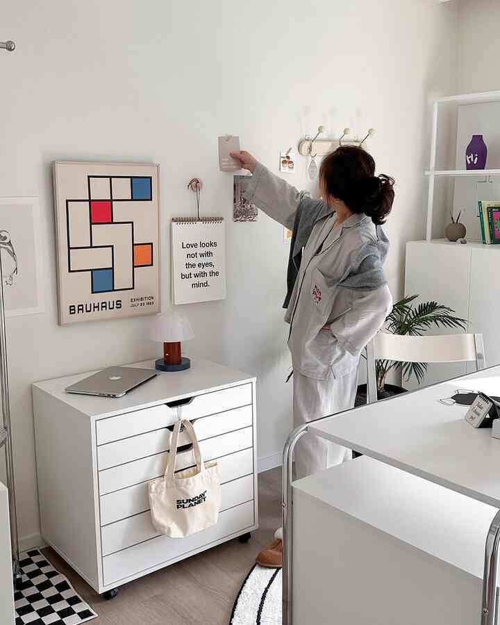 白とナチュラルウッド調のダイニングルームに、本棚とランプ、壁の額縁が配置されたミニマルなホームオフィス空間