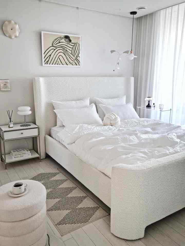 Bright white bedroom featuring a curved bouclé fabric bed with white bedding, a nightstand, and a rug creating a cozy atmosphere