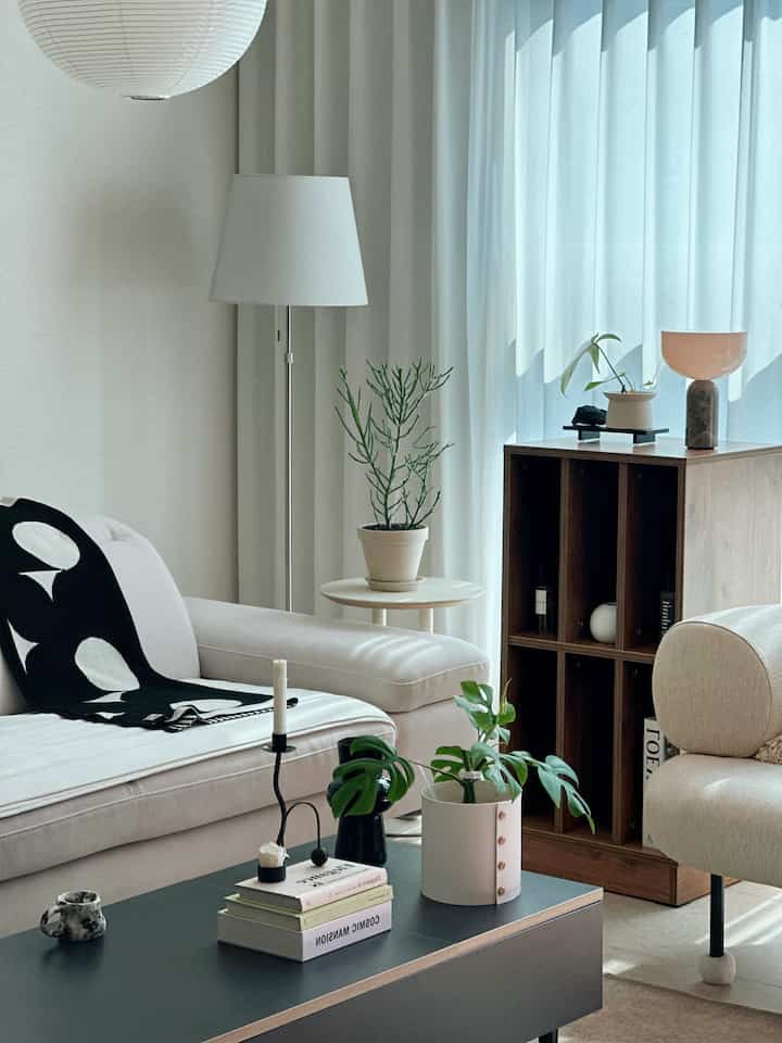 A white and natural toned living room featuring a white sofa, wooden bookshelf, plants, and floor lamp creating a clean atmosphere