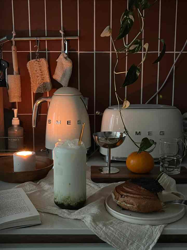 ダークなテラコッタタイルの壁と白いSMEG家電があるキッチン空間、テーブルに置かれたホームカフェの飲み物とデザートが居心地の良い雰囲気を演出