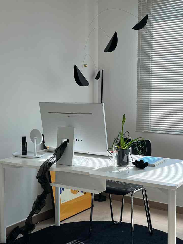 白と黒のトーンの書斎、中央に白いデスクと一体型PC、椅子、窓のアルミニウムブラインドが調和した清潔で暖かみのある空間