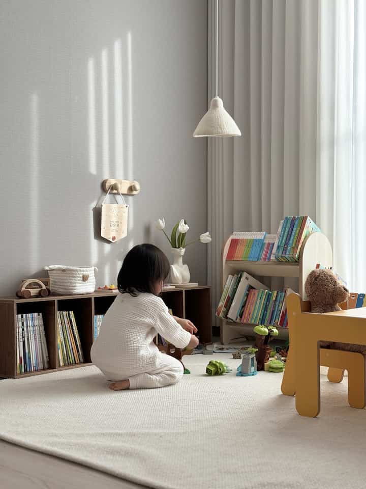 Beige and wood tone kids' room featuring toys, bookshelves, and a cozy play area for a baby under natural light