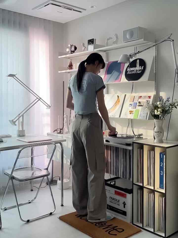 A white and modern small home office featuring a transparent chair and desk, with a woman operating a record player.