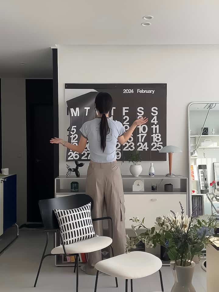 A modern living room in white and black tones featuring a large Stendig Calendar, plants on a cabinet, and a person standing with arms raised