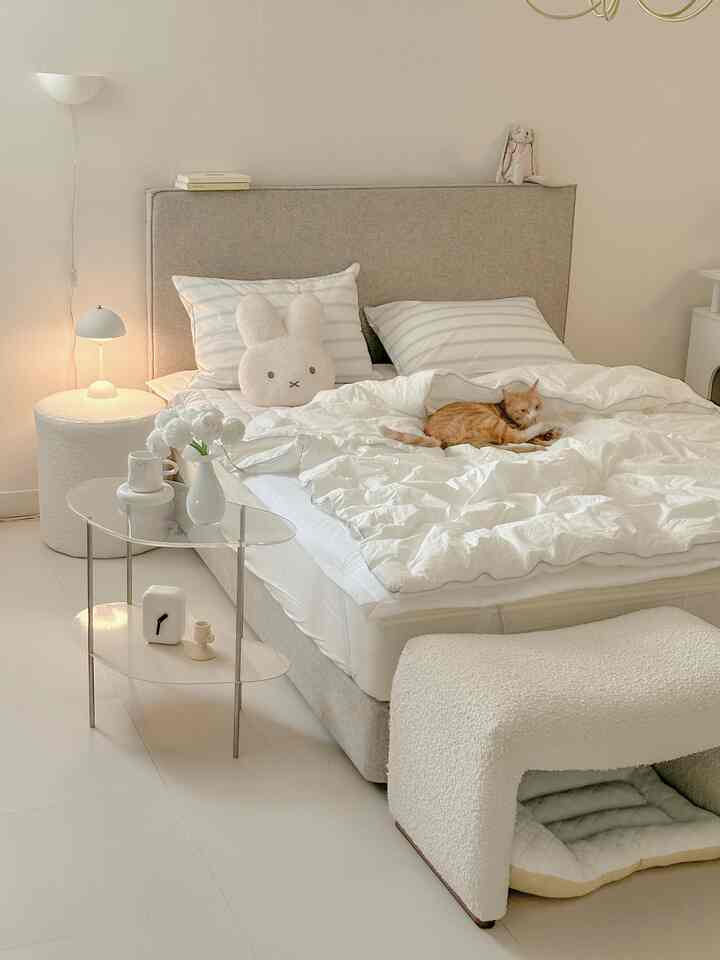 A white and natural-toned bedroom featuring a central bed with a cat, accompanied by a nightstand and table lamp creating a cozy atmosphere