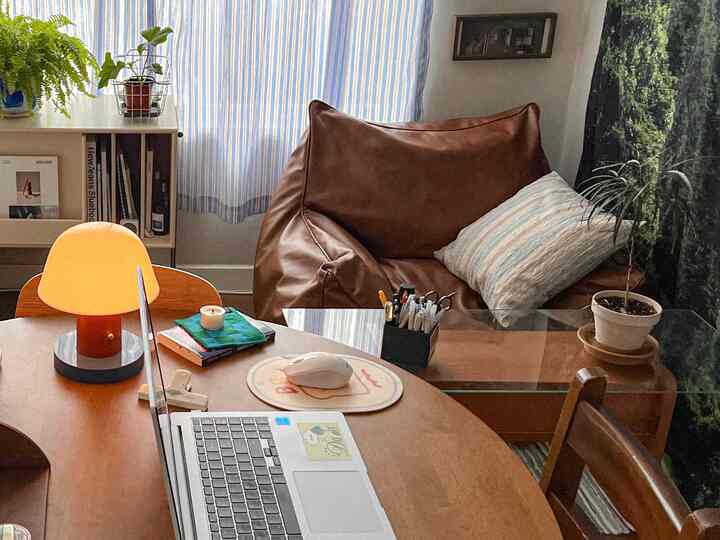 Natural-toned living room featuring a large brown bean bag sofa, glass coffee table, window curtains, and indoor plants creating a cozy atmosphere