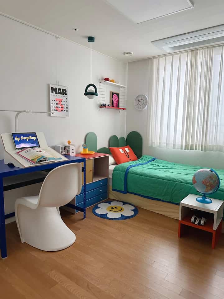 白い壁と木目調の床が際立つ子供部屋、ブルーの机とグリーンの布団がかわいらしく配置された明るく整った空間