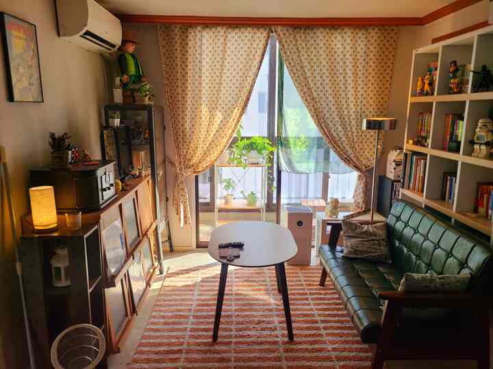 暖かいベージュとブラウンの色調のリビング、グリーンのレザーソファと本棚、柄のカーテンとラグが雰囲気のあるインテリア