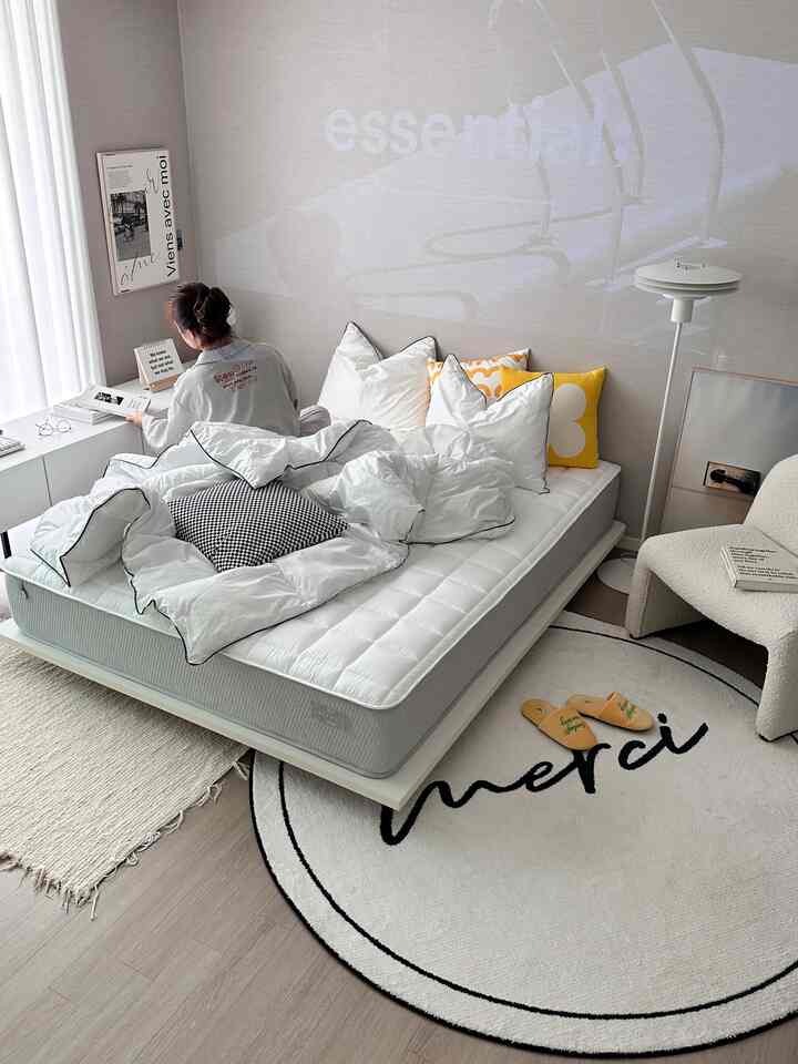 White and beige toned bedroom featuring a low-profile bed frame with mattress, rugs, floor lamp, and armchair in a simple cozy setting