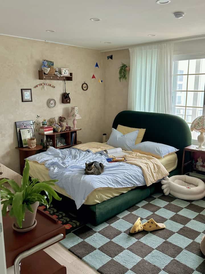 ベージュの壁と木目調のインテリアが調和した寝室、青と黄色の掛け布団を備えたファブリックベッド、中央に猫が座っている印象的な空間