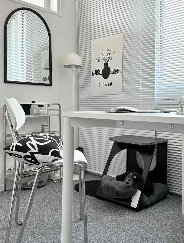 A modern study space in white and gray tones featuring a desk, chair, blinds, and a cozy pet house under the desk