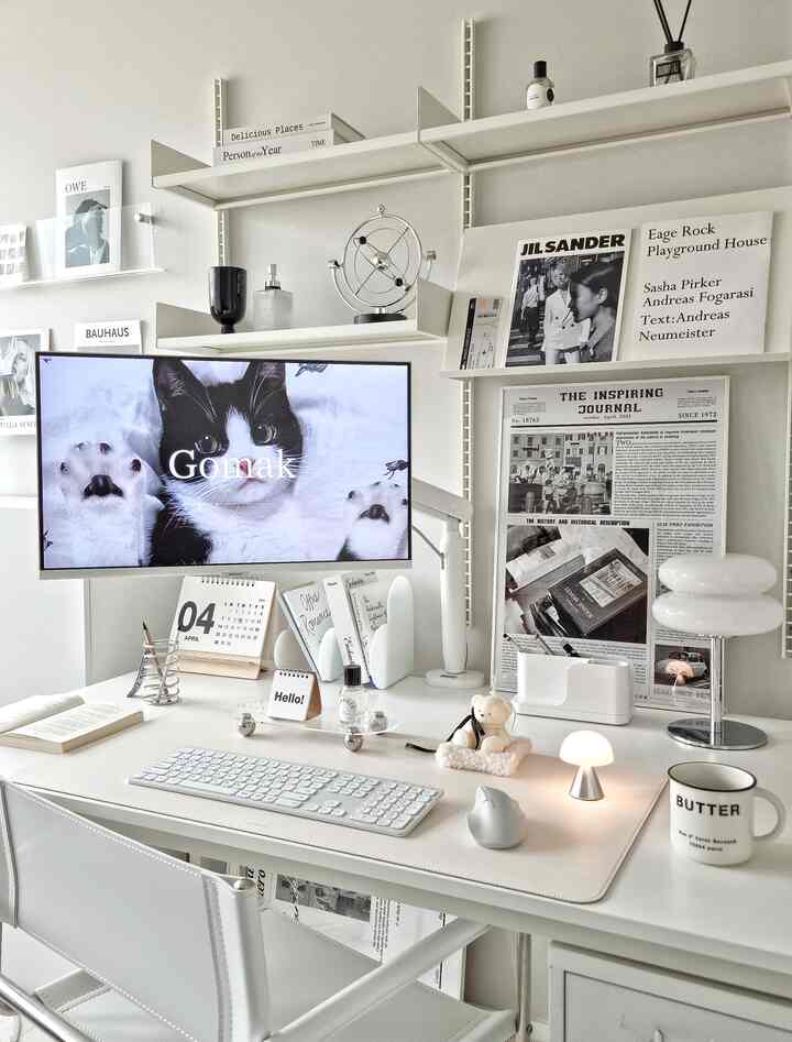 White-toned studio apartment home office featuring a white desk, smart monitor, wall shelves, and minimalist decor creating a sleek workspace