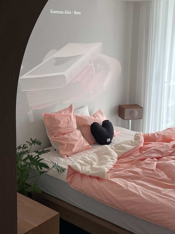 A cozy bedroom with pink and ivory bedding, wood tone bed frame, and white sheer curtains