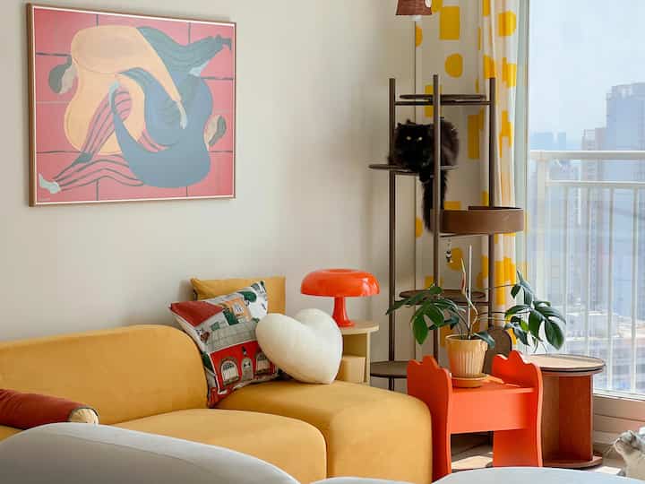 Bright and modern living room featuring a yellow sofa, orange side table, cat tower with a cat, and a plant near large window