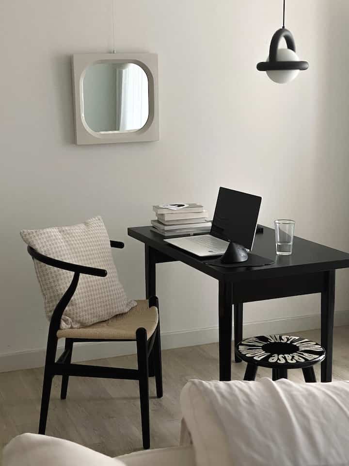 A compact home office space in black and white tones featuring a black desk with laptop and books, a chair with beige cushion in a modern interior
