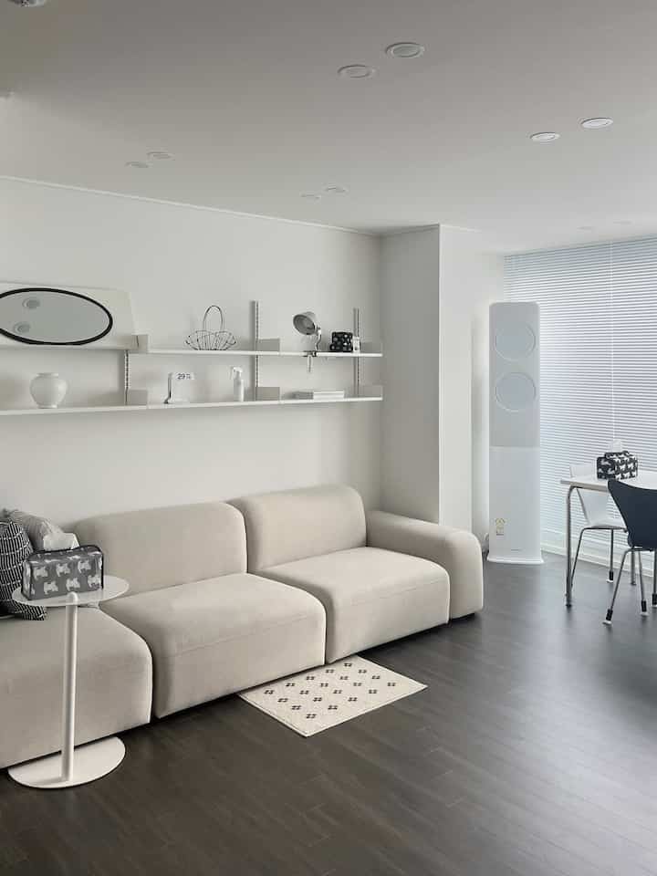 A modern living room in white and brown tones featuring a beige sofa and wall shelving with a clean, minimal atmosphere