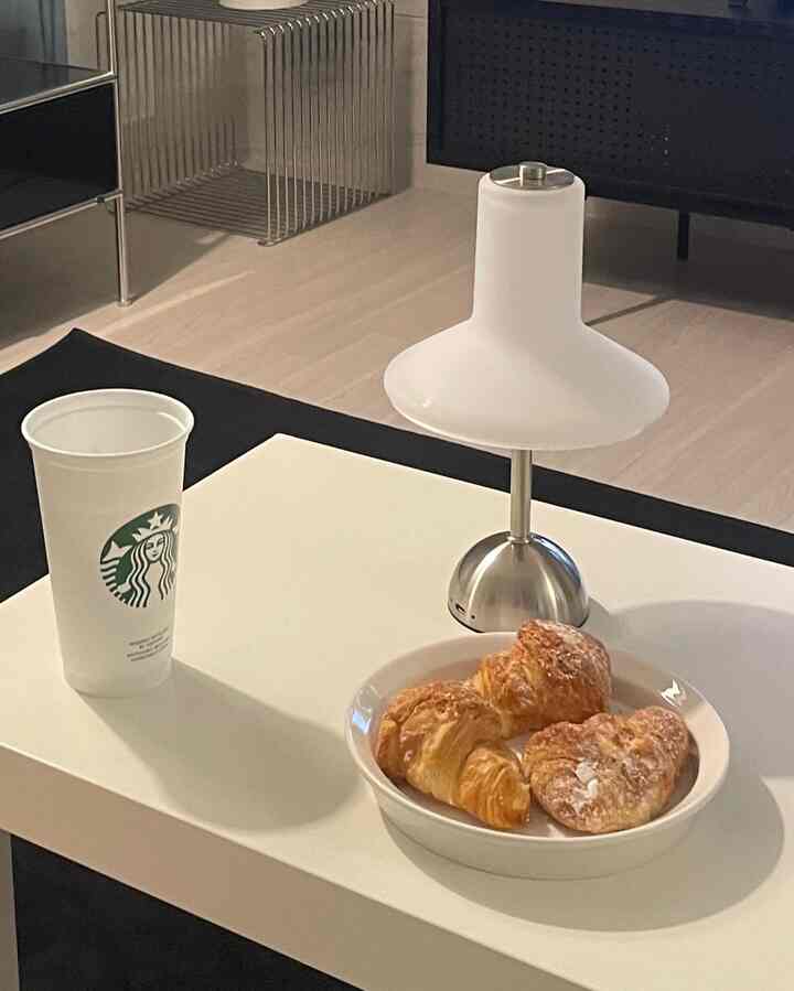 白とシルバーのトーンのリビングルーム、中央に白いコーヒーテーブルとモノルーチェのスタンドランプ、クロワッサンと飲み物が置かれたシンプルなインテリア