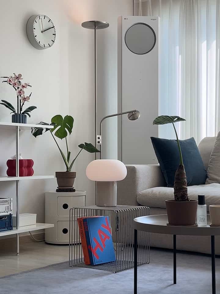 White-toned living room featuring cylindrical storage cabinet, floor lamps, coffee table, and plants in a simple modern setting
