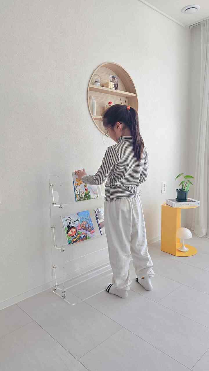White-toned living room featuring a circular wooden shelf, transparent acrylic bookshelf, and a child reading in a simple modern space