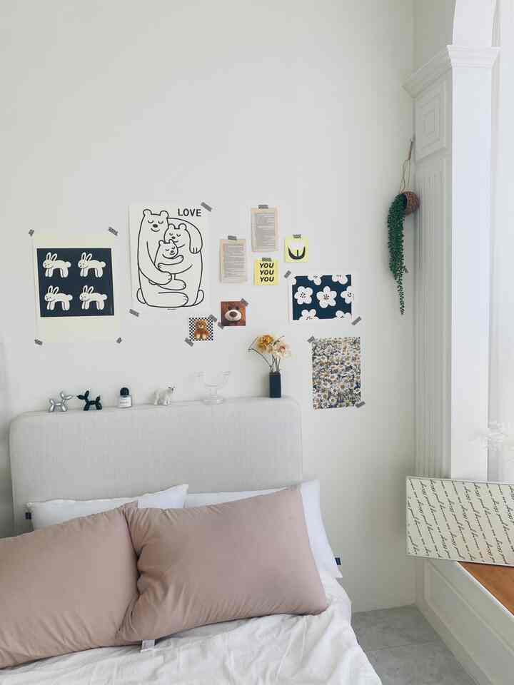 A bedroom with white walls and beige pillows, featuring black-and-white illustrations and floral posters on the wall in a cozy setting