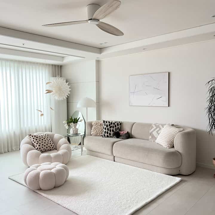 白とベージュの色調のリビング、柔らかな曲線のソファとスツール、クッションが温かみのある雰囲気を演出する空間