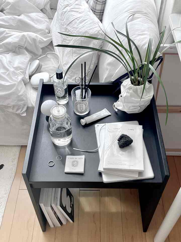 白い寝具と黒いサイドテーブルが配置されたミニマルな寝室、ディフューザーと植物がアクセントの清潔感のあるインテリア