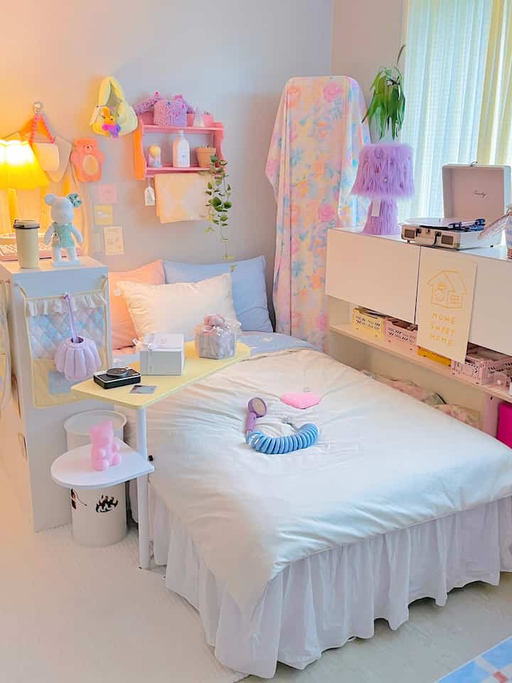 Pastel and white themed small bedroom featuring a central bed with duvet, small bed table, lighting, and decorative objects creating a cozy atmosphere