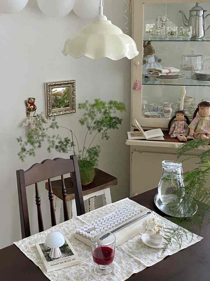 クリーム色のペンダントライトが吊るされたヴィンテージ風のリビング、木製チェアと飾り棚、人形、植物、透明なグラスが置かれた落ち着いた空間