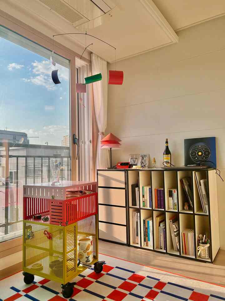 白い壁とバルコニーの扉があるリビング、赤いランプと黄色の収納カートが印象的なモダンなインテリア