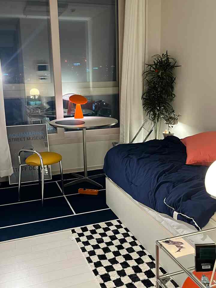 ホワイトとブルーのトーンのワンルーム、オレンジのテーブルランプとナイトテーブル、ラグが配置されたモダンな空間